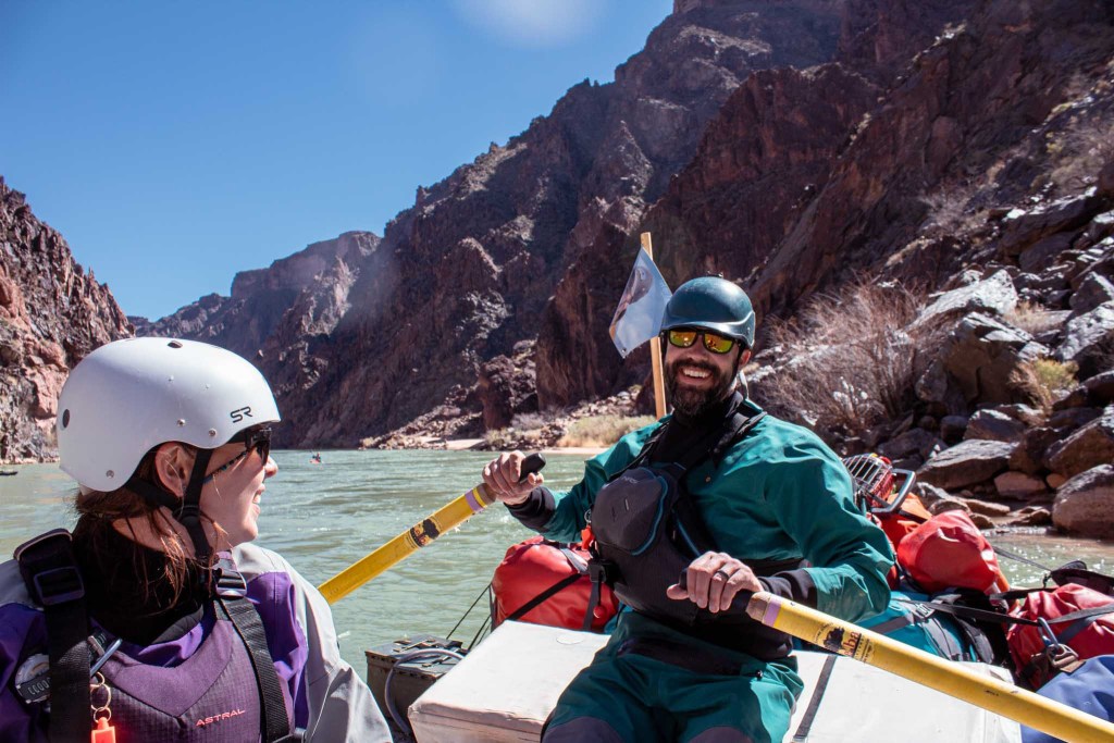 man and woman smiling rowing raft grand canyon in dry suits
